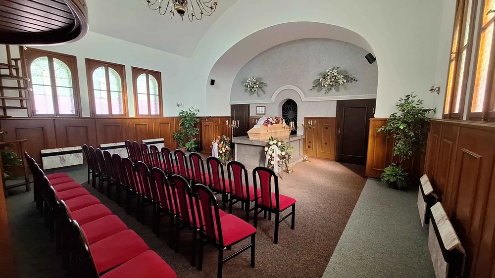 Obřadní síň s klenutým stropem, dřevěným obložením a řadami židlí s červeným čalouněním, světlá rakev s květinami na katafalku, věnce na stojanech a svícny
