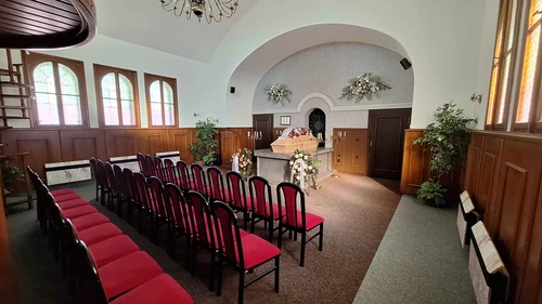 Obřadní síň s klenutým stropem, dřevěným obložením a řadami židlí s červeným čalouněním, světlá rakev s květinami na katafalku, věnce na stojanech a svícny