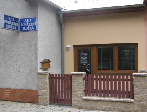 Vchod do provozovny AMI Náplava Zlín pohřební služba s modrými cedulemi na fasádě, prosklenými dveřmi a nízkým dřevěným plotem.
