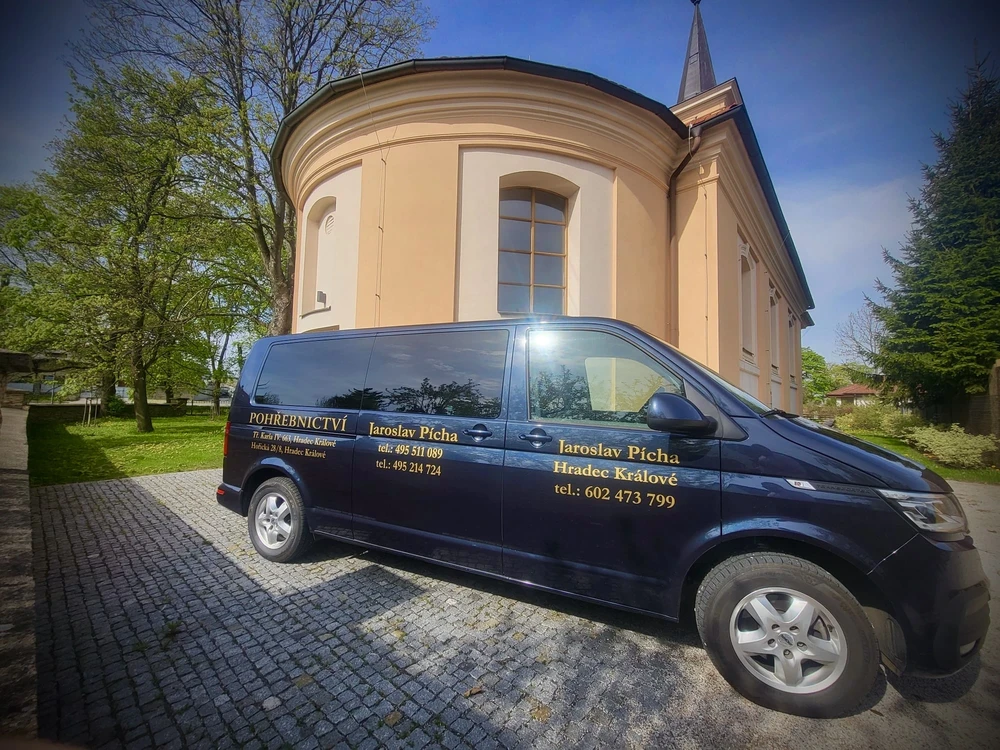 Tmavě modrý pohřební vůz Volkswagen Pohřebnictví Jaroslav Pícha se zlatými nápisy a kontakty před kostelem s kruhovou apsidou v Hradci Králové.