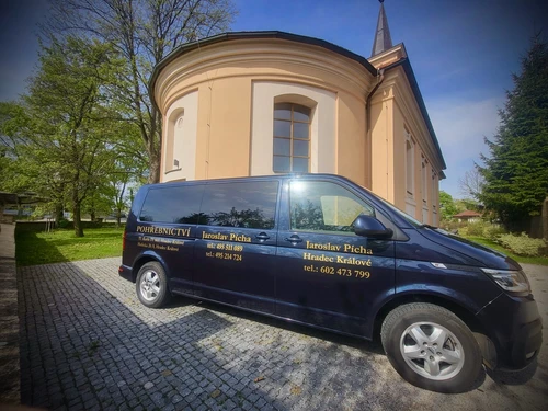 Tmavě modrý pohřební vůz Volkswagen Pohřebnictví Jaroslav Pícha se zlatými nápisy a kontakty před kostelem s kruhovou apsidou v Hradci Králové.