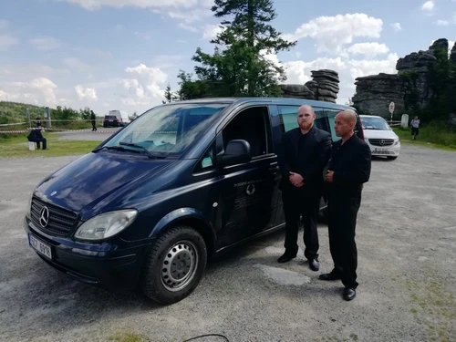 Dva zaměstnanci pohřební služby v černých oblecích u tmavého pohřebního vozu Mercedes-Benz Vito na parkovišti pod pískovcovými skalními útvary.