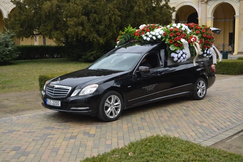 Černý pohřební vůz Mercedes s věnci a květinovými dary na střeše, zaparkovaný před hřbitovní budovou s arkádami.