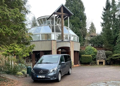 Pohřební vůz Mercedes-Benz Vito před budovou se skleněnou kruhovou nástavbou a dřevěným křížem, obklopenou vzrostlými jehličnany.