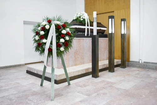 Tmavá rakev na katafalku v obřadní síni, vedle věnec z rudých a bílých růží se stuhami a vysoká smuteční svíce.