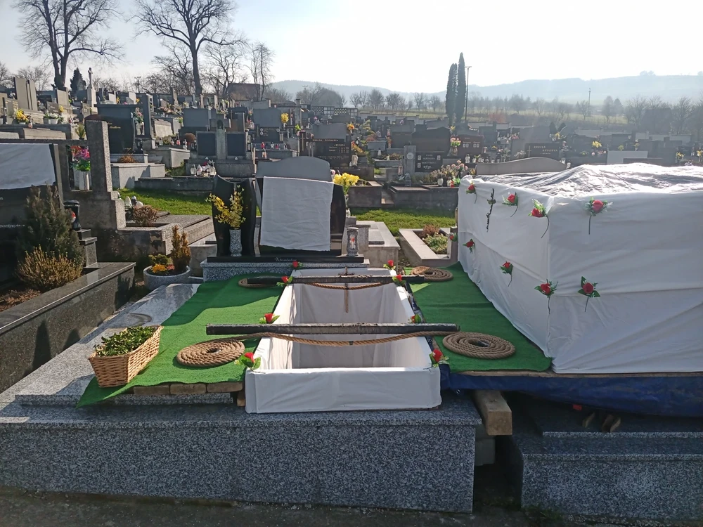 Hrob připravený k uložení, vystlaný bílou tkaninou s růžemi, se spouštěcími lany, břevny a košem s chvojím.