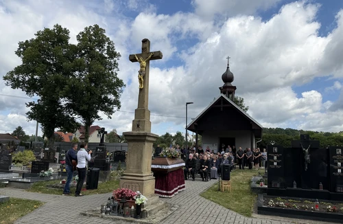 Smuteční obřad na hřbitově u kamenného kříže, rakev s květinami, hudebníci s kytarou a shromáždění hosté před kaplí.