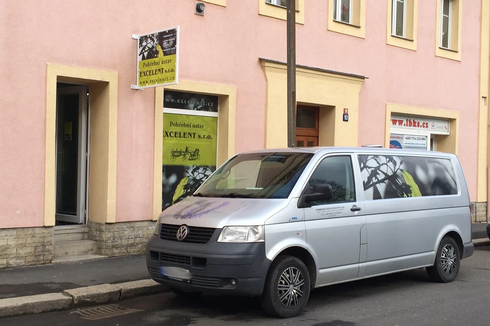 Provozovna Pohřebního ústavu EXCELENT s cedulí na fasádě a stříbrným pohřebním vozem Volkswagen Transporter zaparkovaným před vchodem.