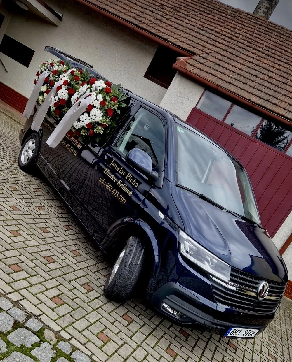 Tmavě modrý pohřební vůz Volkswagen Pohřebnictví Jaroslav Pícha s otevřenými zadními dveřmi a smutečními věnci z červených růží a bílých chryzantém.