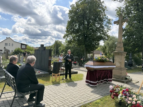 Řečník pohřební služby přednáší smuteční řeč u mikrofonu, vedle rakev s květinovou vazbou u kamenného kříže.