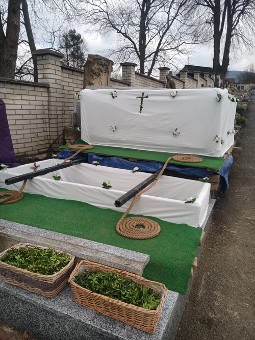 Připravený hrob s bílým dekorem a krucifixem, vedle zakrytá vykopaná hlína a proutěné koše s chvojím k zasypání.