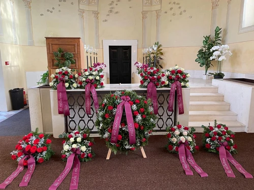 Smuteční věnce a kytice z červených a bílých růží s vínovými stuhami u katafalku v obřadní síni se svícny a orchidejemi.