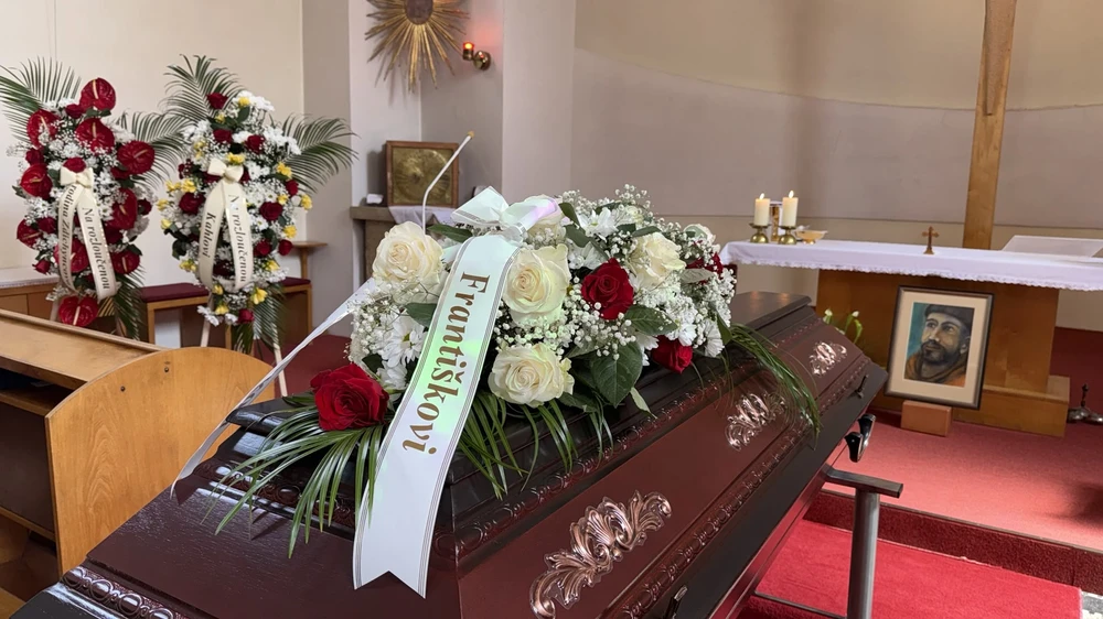 Tmavá rakev s bílo-červenou kyticí z růží v kostele, v pozadí oltář s hořícími svícemi a dvě smuteční kytice na stojanech.