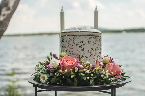 Bílá porcelánová urna s květinovým dekorem a svícemi, obklopená růžemi a eustomami na stolku u hladiny jezera.