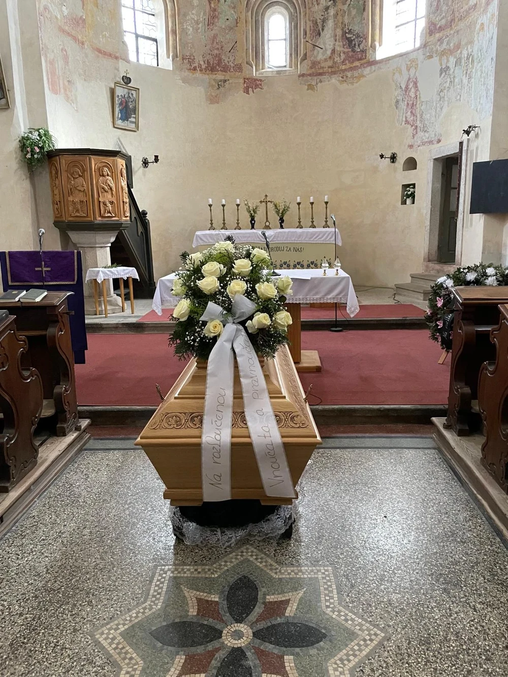 Světlá dřevěná rakev s kyticí bílých růží a bílou stuhou v kostele s nástěnnými malbami, oltářem se svícny a vyřezávanou kazatelnou.