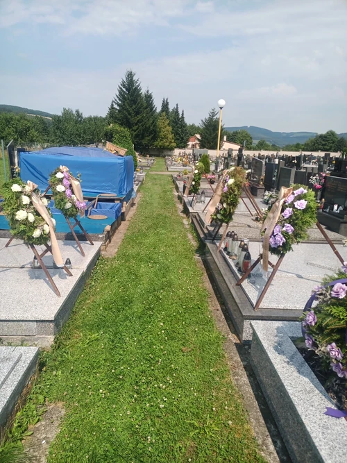 Připravený hrob na vesnickém hřbitově, hlína zakrytá modrou plachtou, věnce s bílými a fialovými růžemi na stojanech.