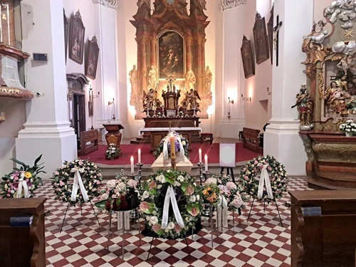 Smuteční obřad v barokním kostele, rakev se svící a hortenziemi před oltářem, věnce z růží a toulitek na stojanech, svíce a šachovnicová podlaha