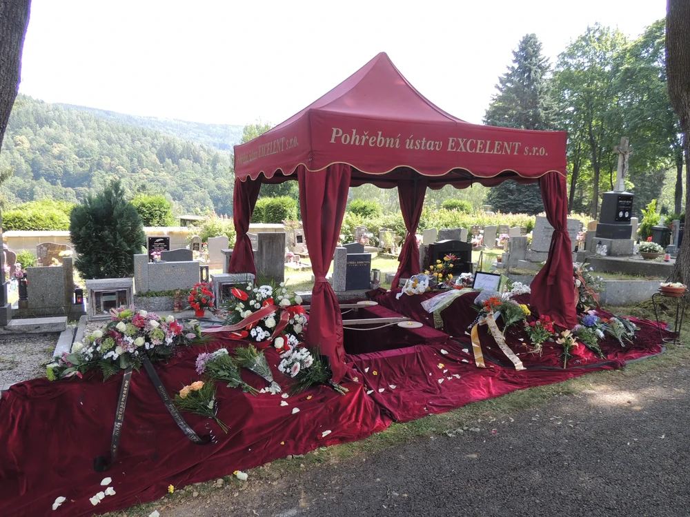 Vínový stan Pohřebního ústavu EXCELENT nad otevřeným hrobem, okolí pokryté sametovou látkou se smutečními kyticemi a věnci, v pozadí zalesněné kopce.