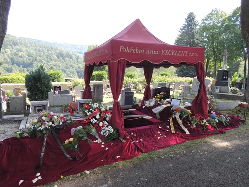 Vínový stan Pohřebního ústavu EXCELENT nad otevřeným hrobem, okolí pokryté sametovou látkou se smutečními kyticemi a věnci, v pozadí zalesněné kopce.