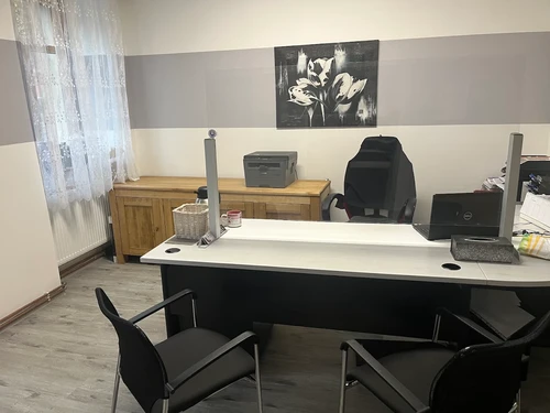 Kancelář pohřební služby s pracovním stolem, židlemi pro klienty, tiskárnou a komodou u okna.