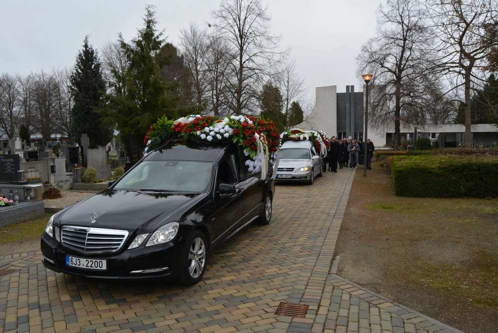 Pohřební průvod přijíždí po dlážděné cestě na hřbitov, v čele vůz s květinovou výzdobou, za ním smuteční hosté.