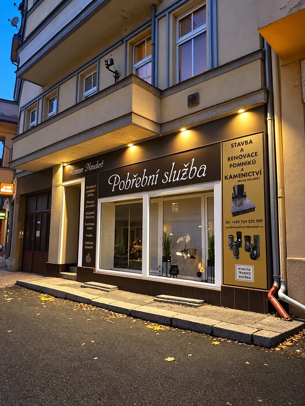 Prosvětlená provozovna pohřební služby Roman Neudert za soumraku, s výlohou a výčtem nabízených služeb.
