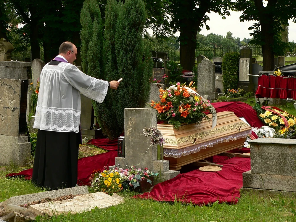 Kněz v rochetě a fialové štole žehná dřevěnou rakev s kyticí oranžových lilií a karafiátů u hrobu na hřbitově.