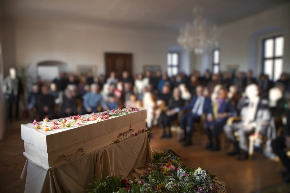 Světlá dřevěná rakev s růžemi v sále plném truchlících, u paty rakve pestrá vazba z lučních květin.