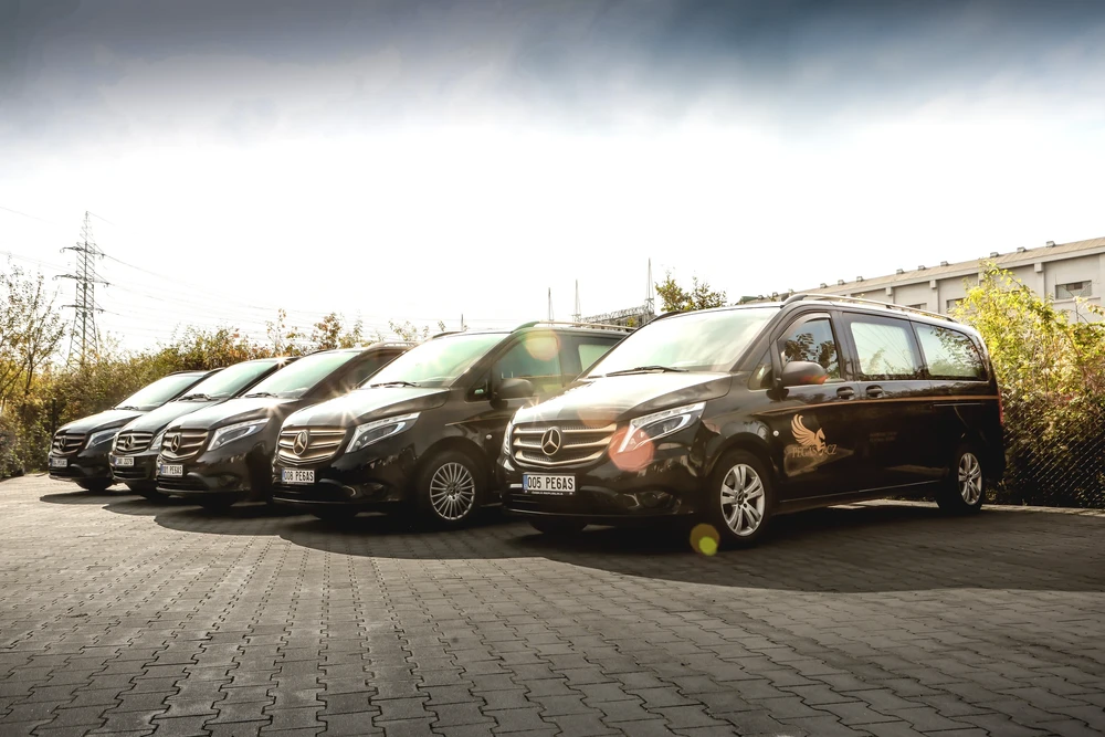 Řada pěti černých pohřebních vozů Mercedes-Benz Vito Pohřební služby Pegas se zlatým logem na parkovišti.