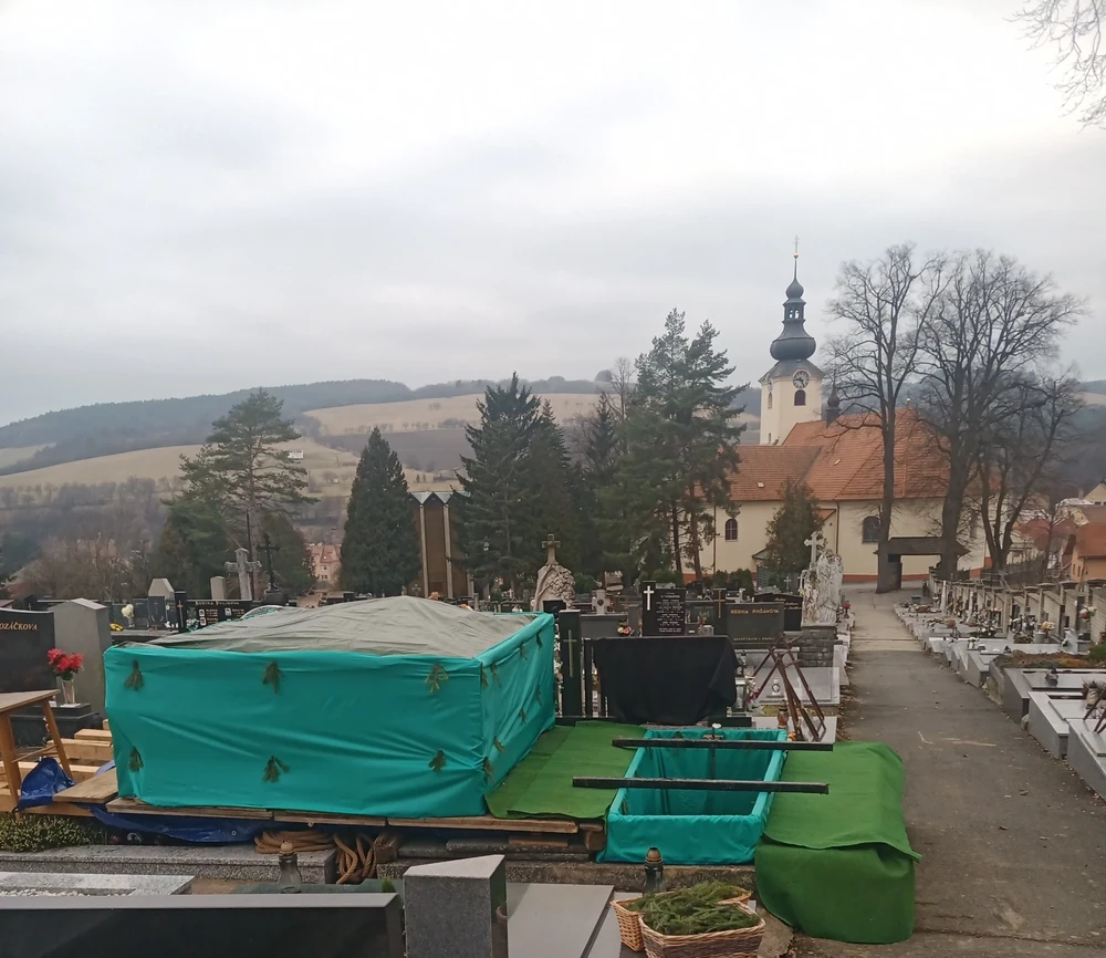 Připravený hrob na hřbitově pod kostelem, hlína zakrytá zelenou plachtou s chvojím, v pozadí zvlněná krajina.