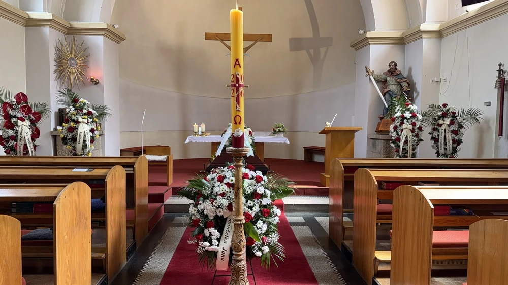 Kostel připravený k pohřbu – svíce s křížem, rakev s kyticí v hlavní uličce a kytice z červených růží u stěn.