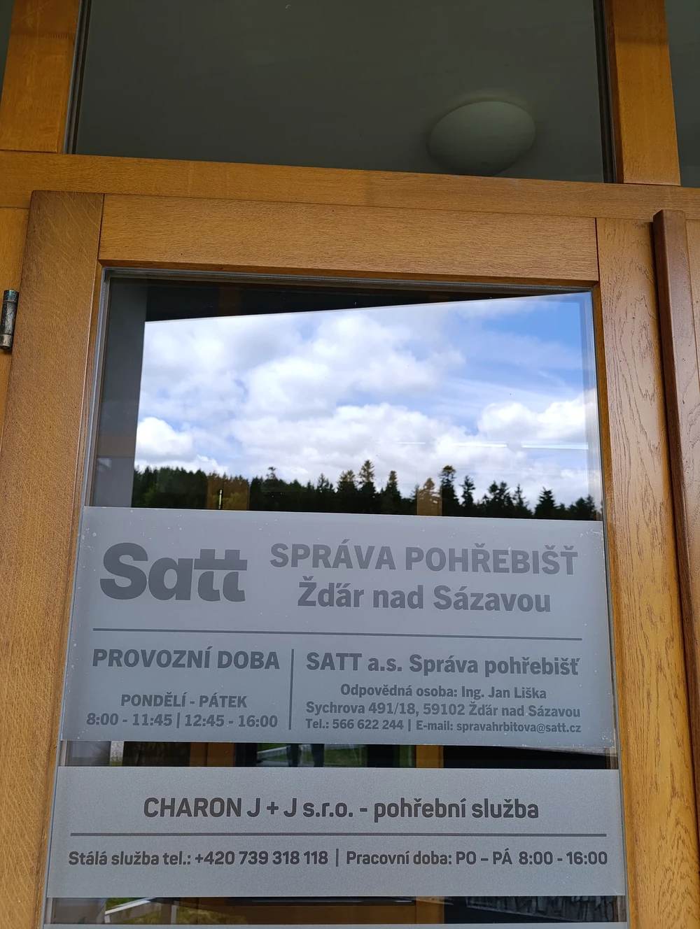 Informační cedule na dveřích: správa pohřebišť Žďár nad Sázavou a pohřební služba Charon J+J s provozní dobou.