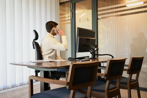 Kancelář pohřební služby HELFI, zaměstnanec telefonuje u pracovního stolu s monitorem, dřevěné židle pro klienty a logo HELFI na skleněných dveřích