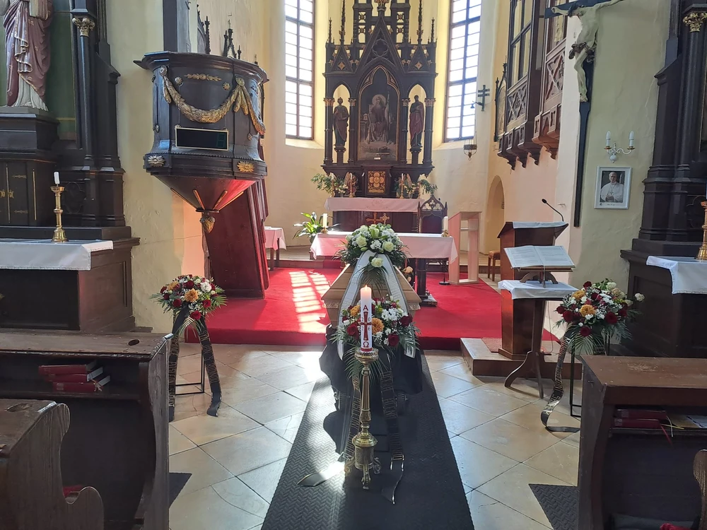 Interiér gotického kostela s rakví v hlavní uličce, velikonoční svící a dvěma vínovými věnci, sluneční pruhy dopadají na červený koberec.
