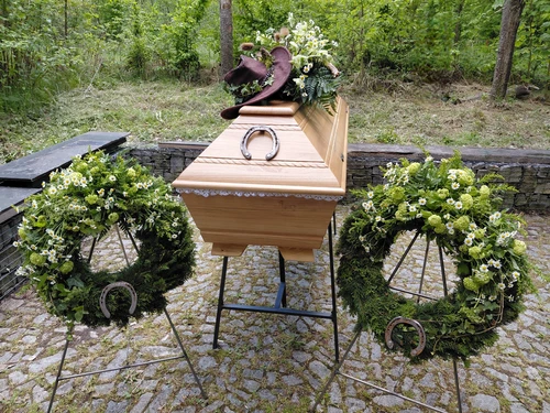 Světlá dřevěná rakev s mysliveckým kloboukem, podkovou a polními květinami při rozloučení v přírodě, po stranách dva věnce s kopretinami a podkovami.