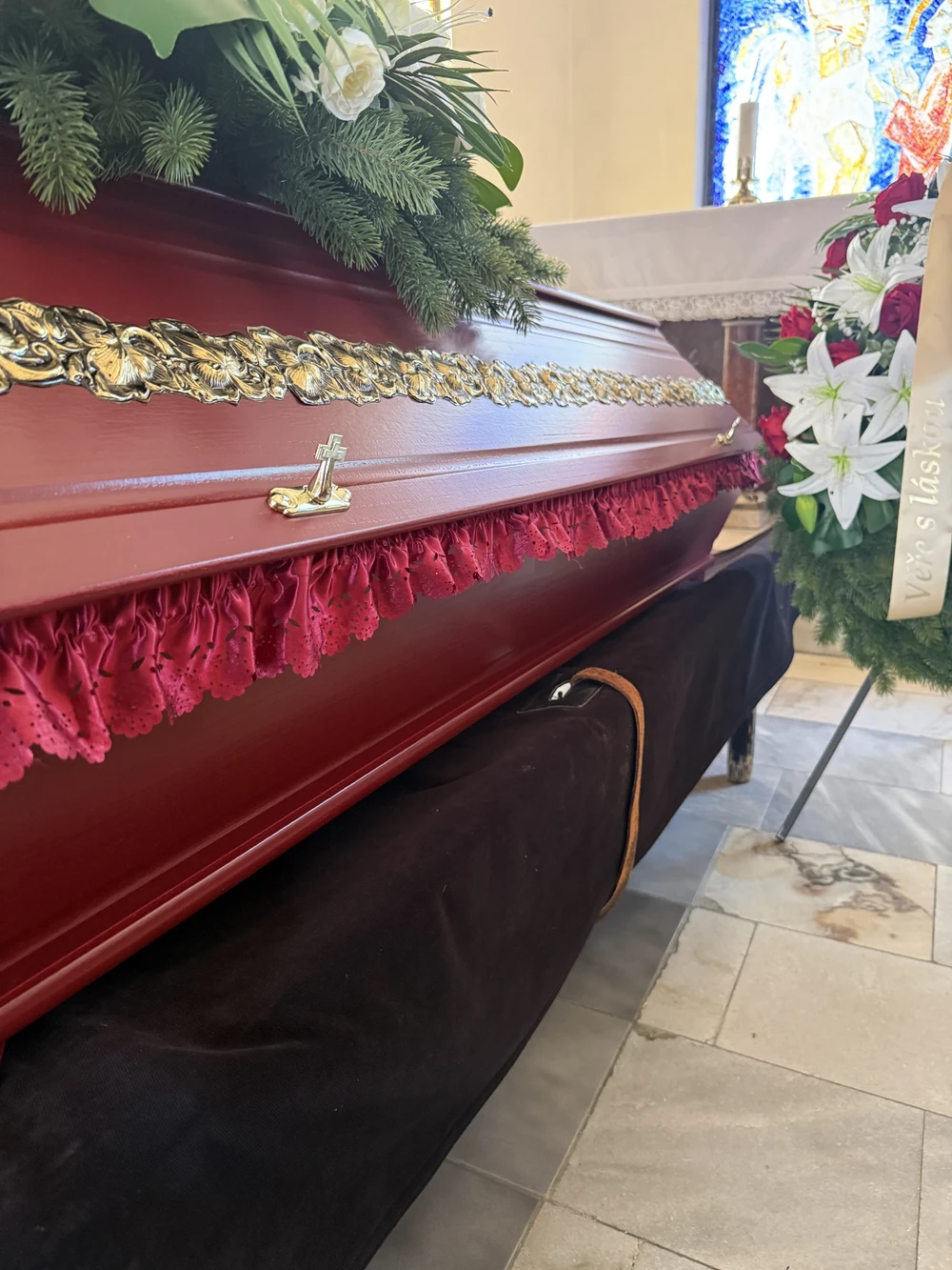 Detail vínové rakve se zlaceným zdobením a řaseným sametovým lemem na tmavě potaženém katafalku, v pozadí vitrážové okno.