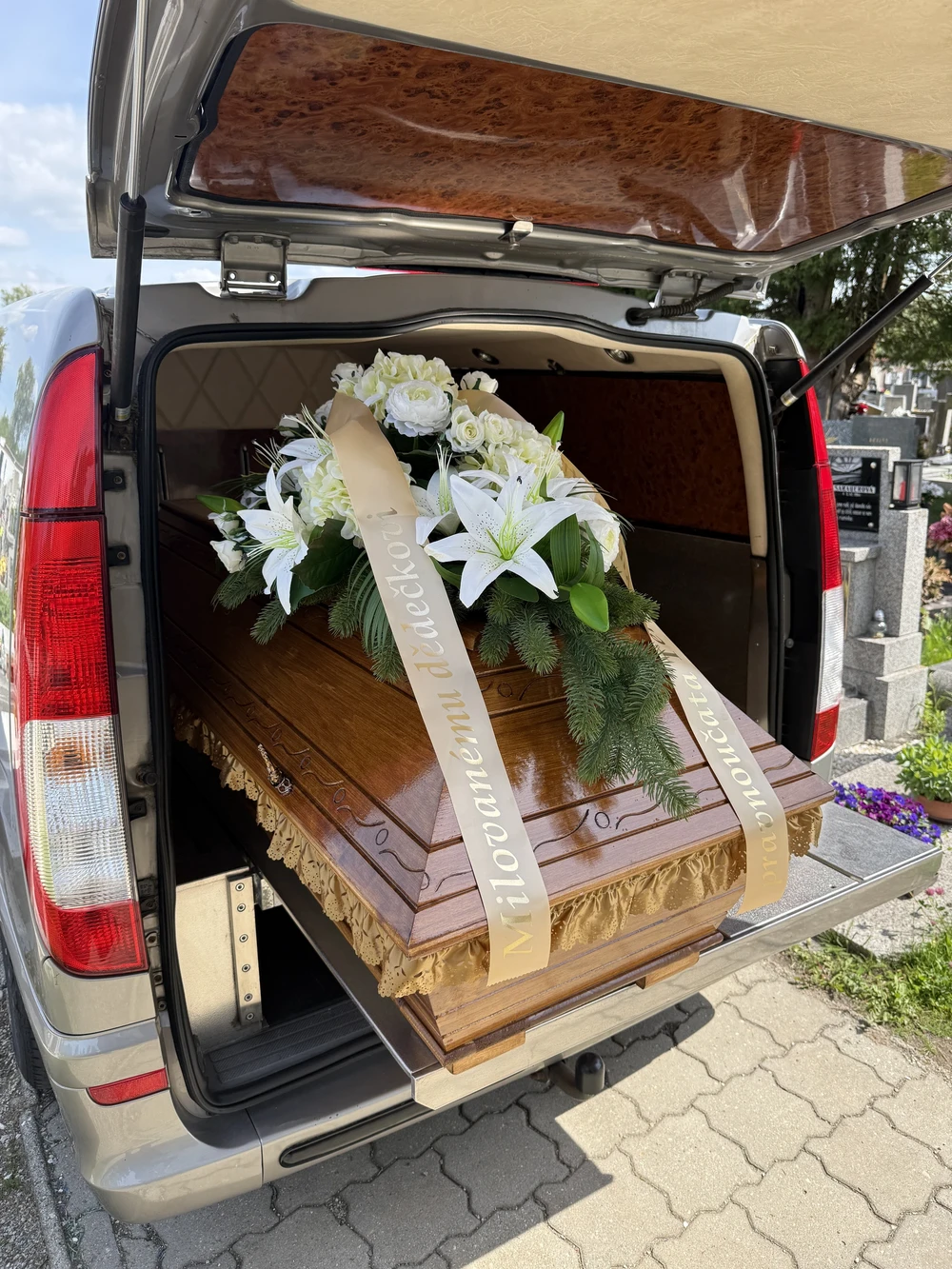 Dřevěná rakev s kyticí z bílých lilií a růží uložená v pohřebním vozu s otevřenými zadními dveřmi, v pozadí hřbitov.